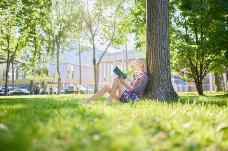Young women sits on grass in park and reads book with warm sunlight filtering through the trees
