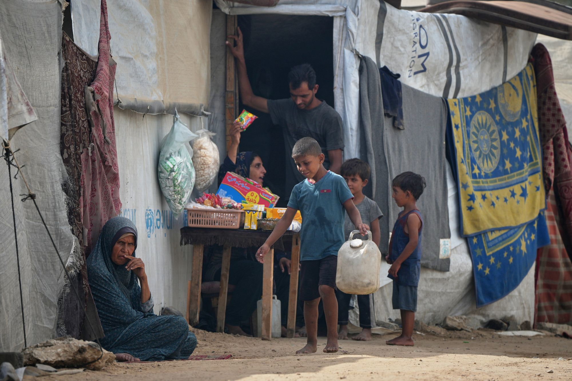 A woman tends her stall as a boy walks by carrying a jerrycan at a tent camp for displaced Palestinians in Deir al-Balah, in the central Gaza Strip