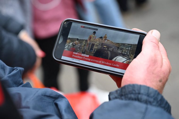 A person watches live BBC news footage of the Carriage procession of US President Donald Trump and Britain's King Charles III travelling in the grounds of Windsor Castle, as they stand in Windsor, on September 17, 2025, during US President Donald Trump's second State Visit to the UK. US President Donald Trump arrived in Britain on Tuesday for an unprecedented second state visit, with the UK government rolling out a royal red carpet welcome to win over the mercurial leader. (Photo by Paul ELLIS / AFP) (Photo by PAUL ELLIS/AFP via Getty Images)