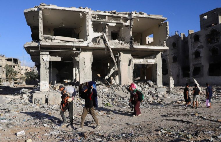 Palestinians walk carrying their belongings past a damaged building in a neighborhood in the aftermath of an Israeli operation, after Israel's government ratified a ceasefire with Hamas, in Gaza City October 10, 2025. REUTERS/ Ebrahim Hajjaj